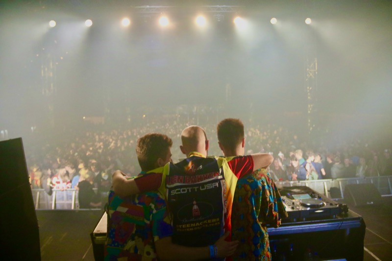 De drie leden van Let's Dance met de armen om elkaar heen, met de rug naar de camera, kijkend naar een vol festivalpubliek