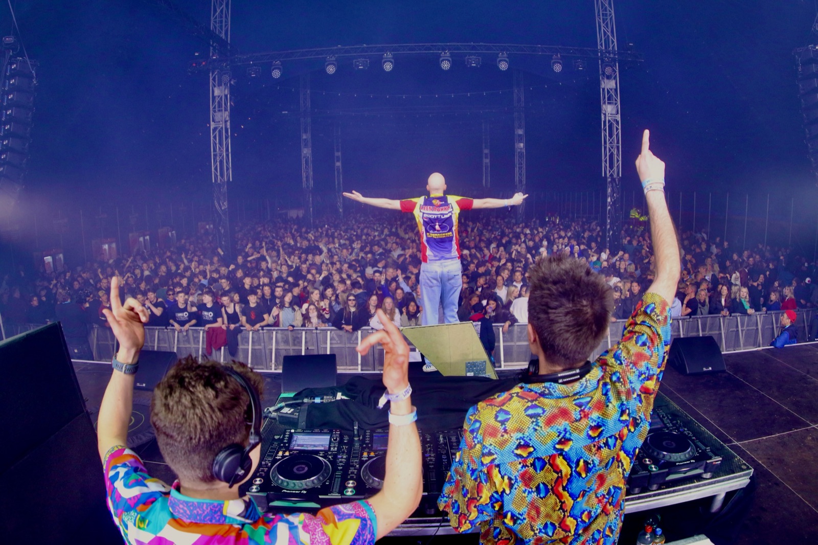 Twee dj's van Let's Dance in kleurrijke hemden met de armen in de lucht achter de draaitafels, MC Tukobino voor een volle festivaltent
